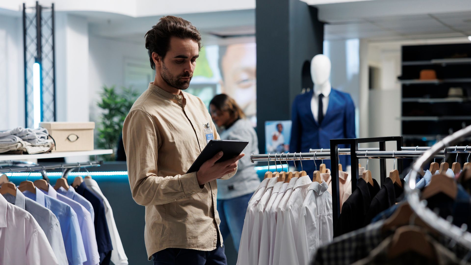 Retail employee using a digital tablet while checking clothing inventory in a modern store with digital displays